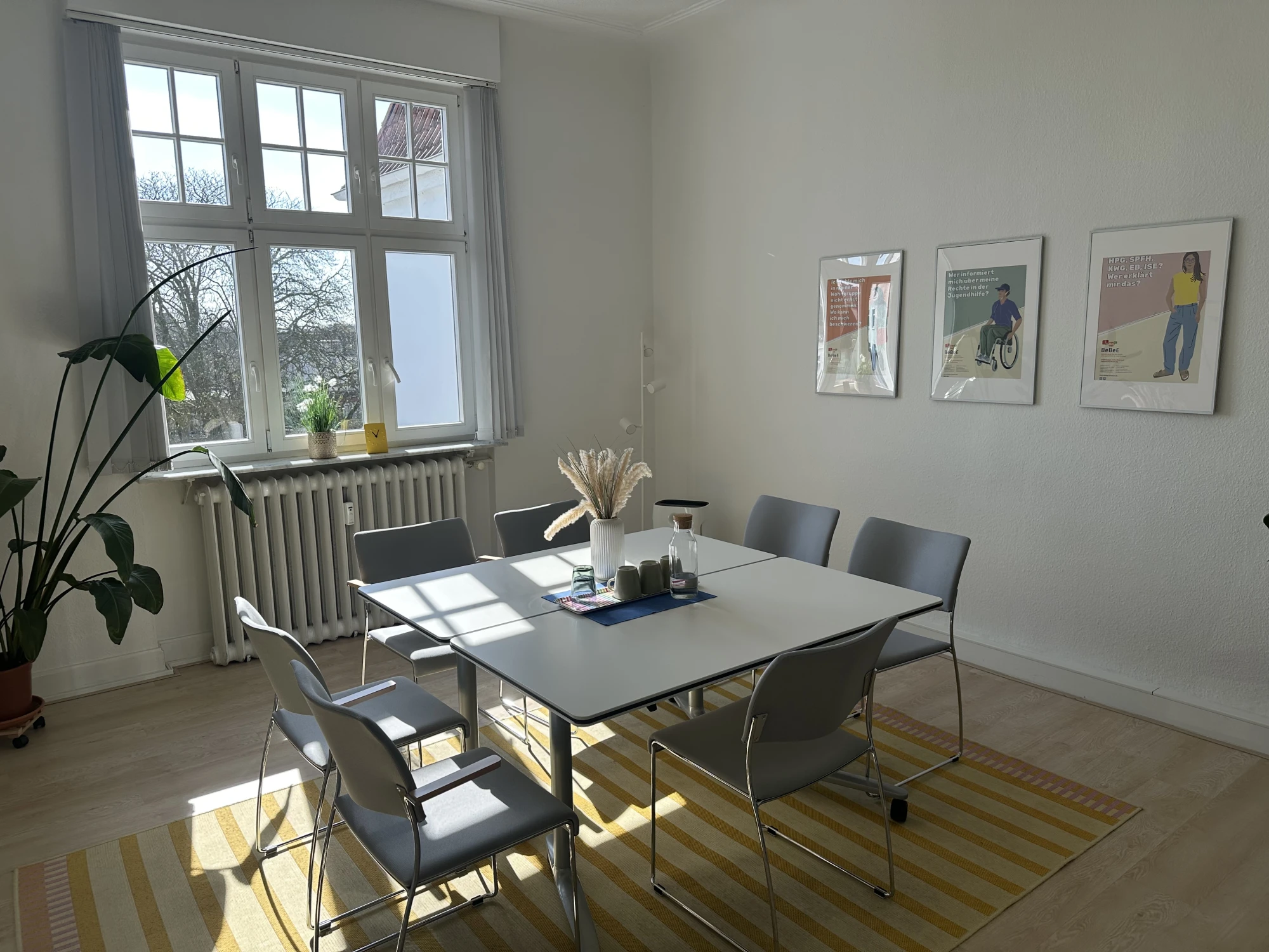 Ein Tisch mit 7 St&uuml;hlen im lichtdurchflutete Besprechungsraum, daneben eine Zimmerpflanze, an der Wand die Illustrationen von BeBeE